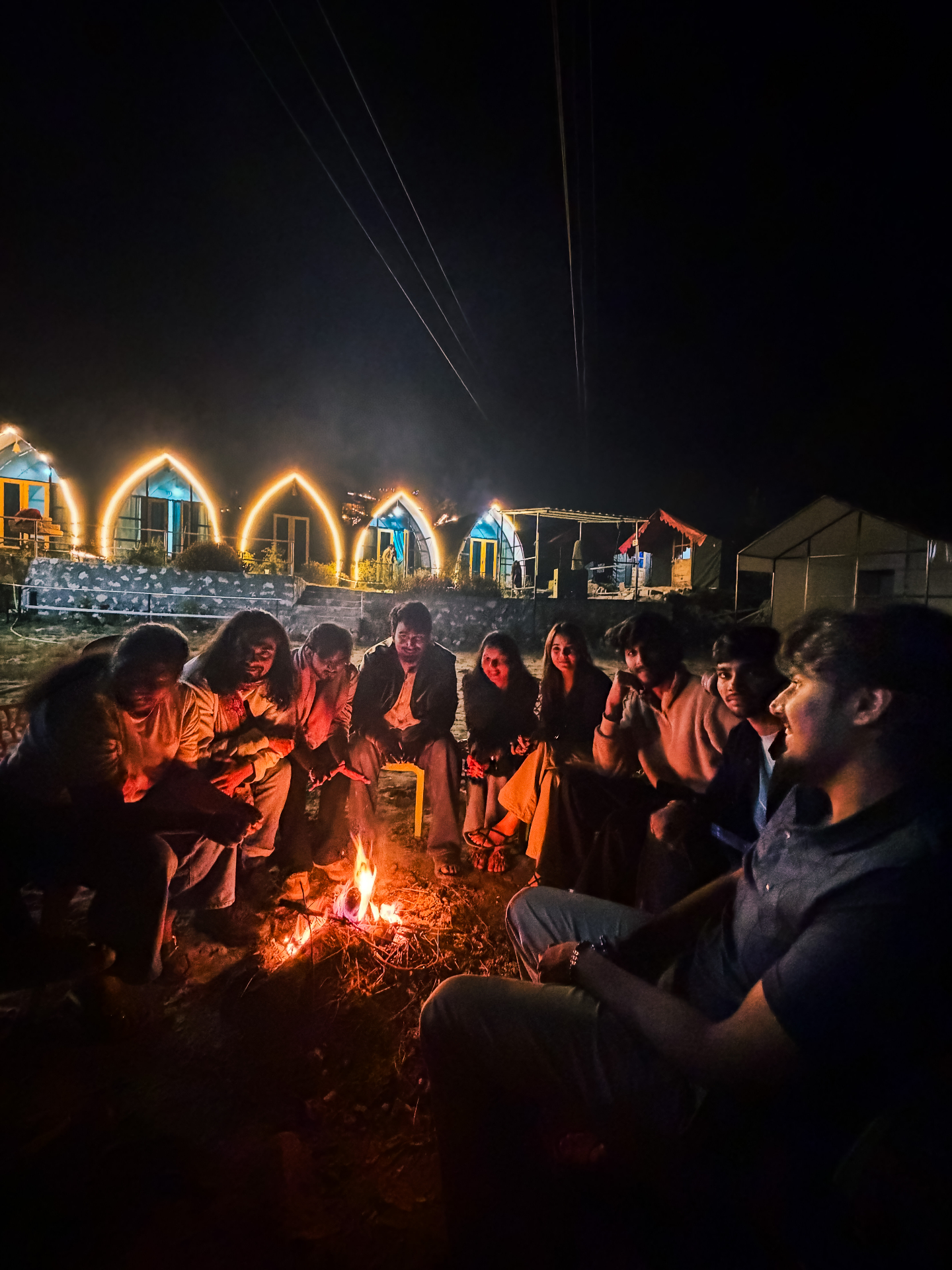 Travellers around a bonfire at night in front of lit mountain cottages